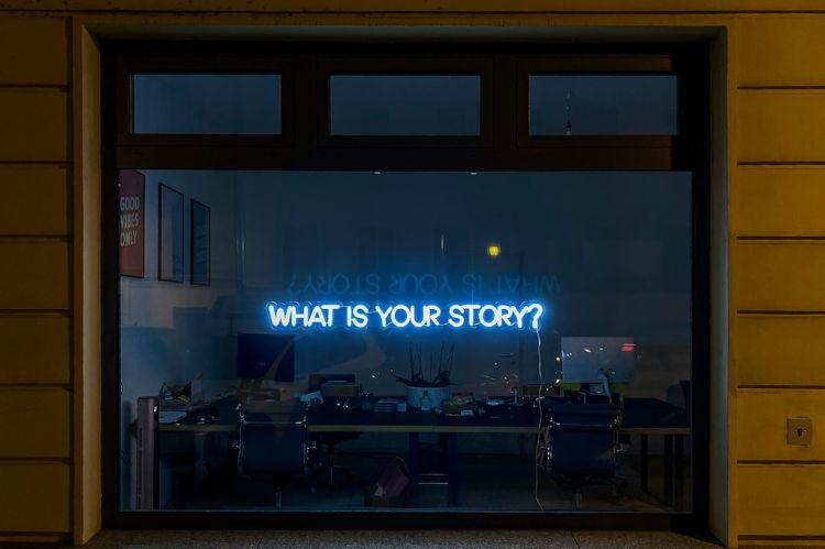 a lit neon window sign displaying the phrase "What is your story?"