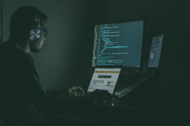 A man sitting in a dark corner with two laptops and a desktop screen.