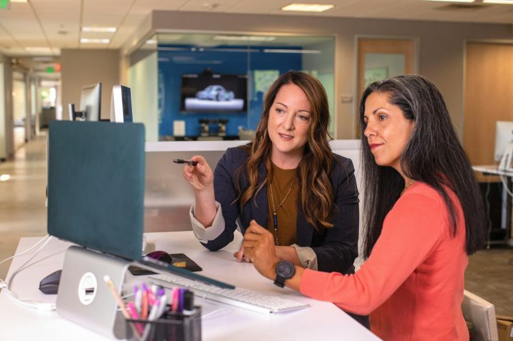 Two business women sitting at a desk and looking at a laptop.