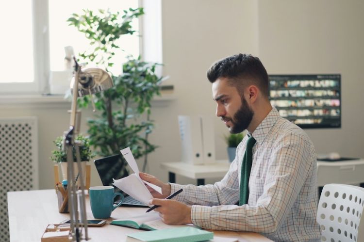 a businessman sitting at a desk reading several papers that he is holding in his hand