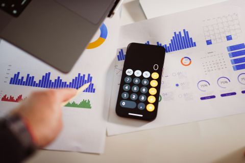 An iPhone calculator sitting next to a laptop on top of charts and diagrams.