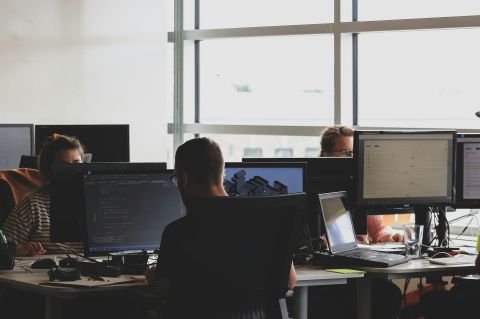 Employees in an open office space sitting on chairs in front of computer monitors.